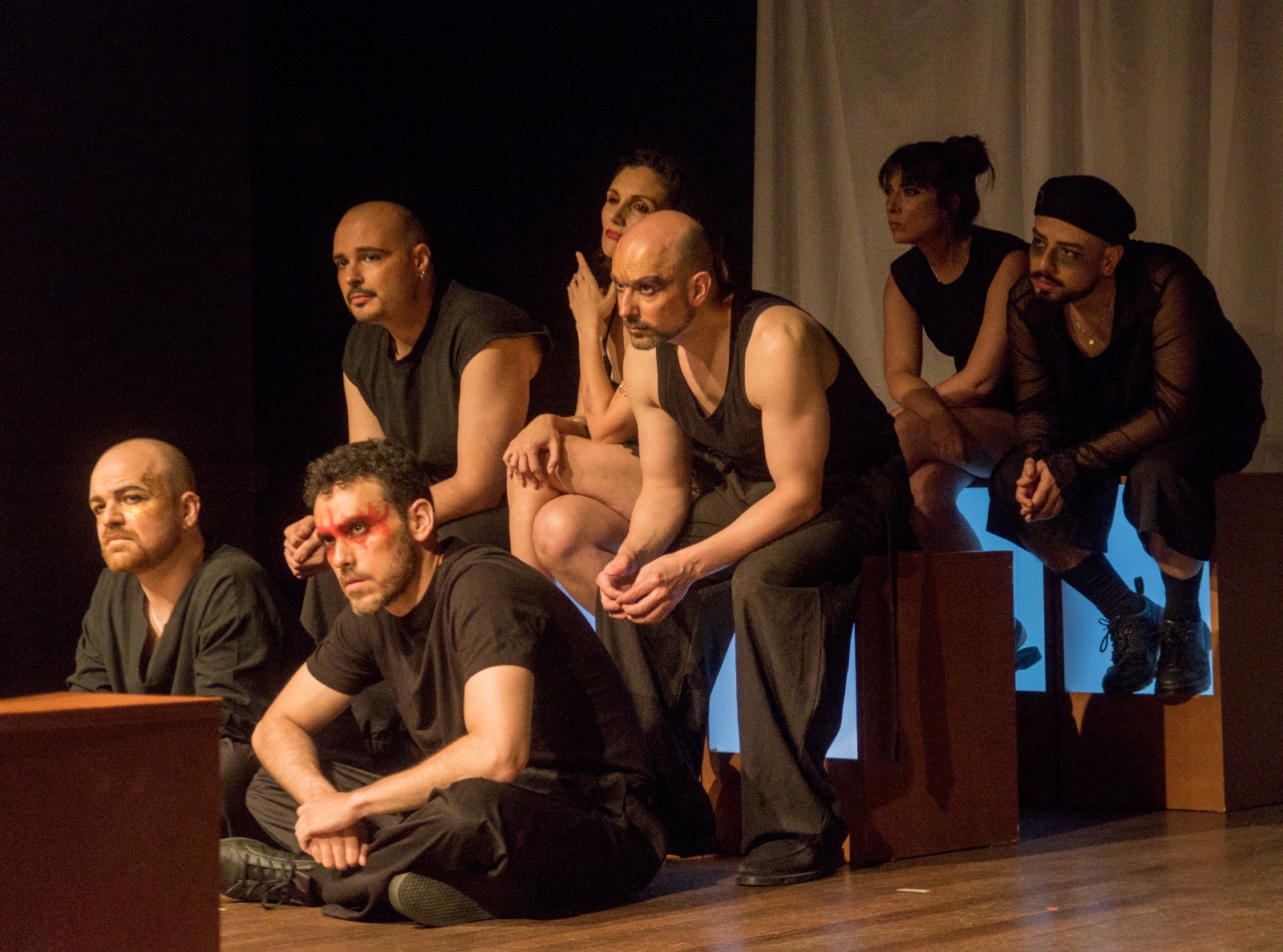 El elenco, vestido de negro, permanece sentado a diferentes alturas sobre el escenario.