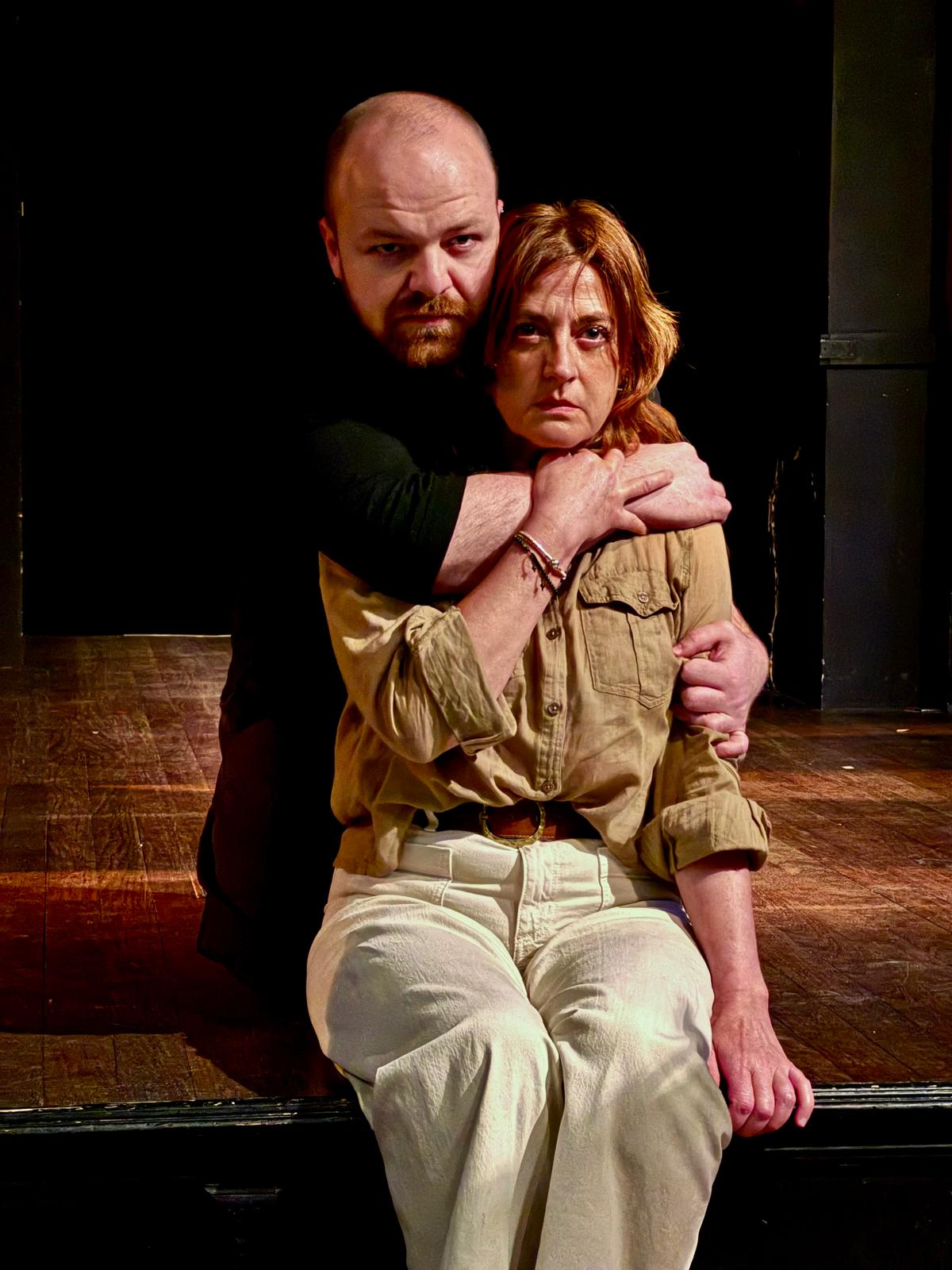 Un actor abraza por los hombros a una actriz sentada en el borde del escenario.